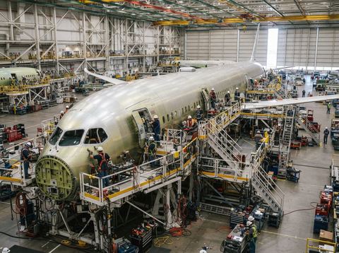 A commercial jet fuselage under construction inside a large, brightly lit aircraft assembly hangar.