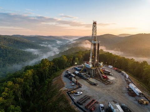 A natural gas drilling rig set against wooded Appalachian hills at sunrise.
