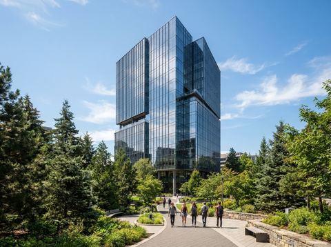 Modern glass office tower on a landscaped corporate campus under a bright blue sky.