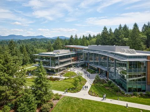 Modern glass office campus building surrounded by evergreen trees under a bright sky, representing a major American software company headquarters.