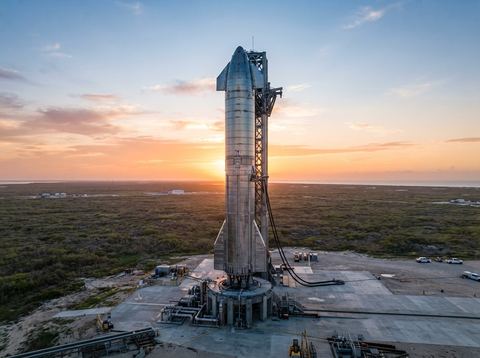 A reusable rocket booster standing on a launch pad in an open Texas landscape at sunrise.