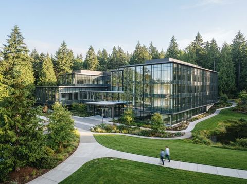 A modern glass corporate office campus building surrounded by trees under bright daylight.